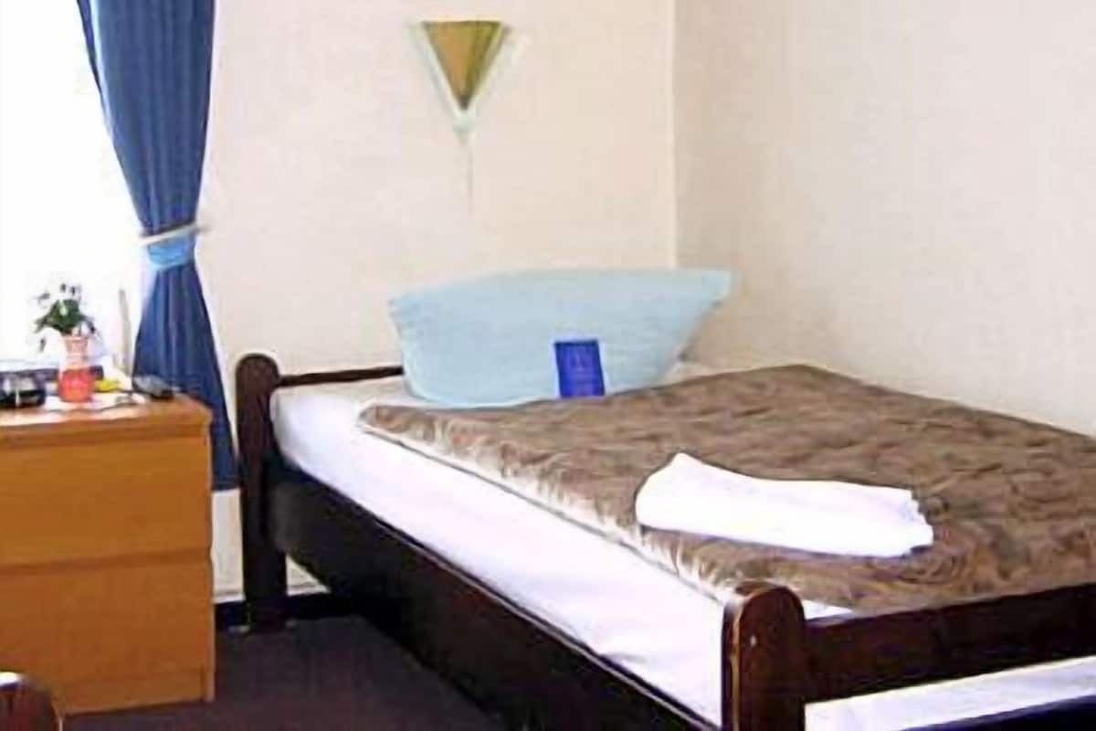 Une petite chambre avec un lit simple, un cadre en bois, un oreiller bleu, un matelas blanc et une table de chevet avec une lampe.