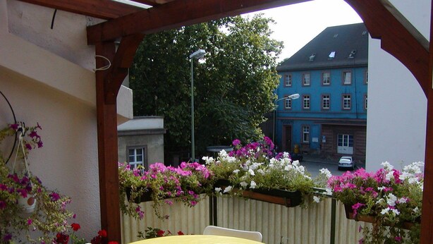 Een ronde tafel met een gele tafelkleed en witte stoelen, omgeven door een houten pergola met bloembedden.