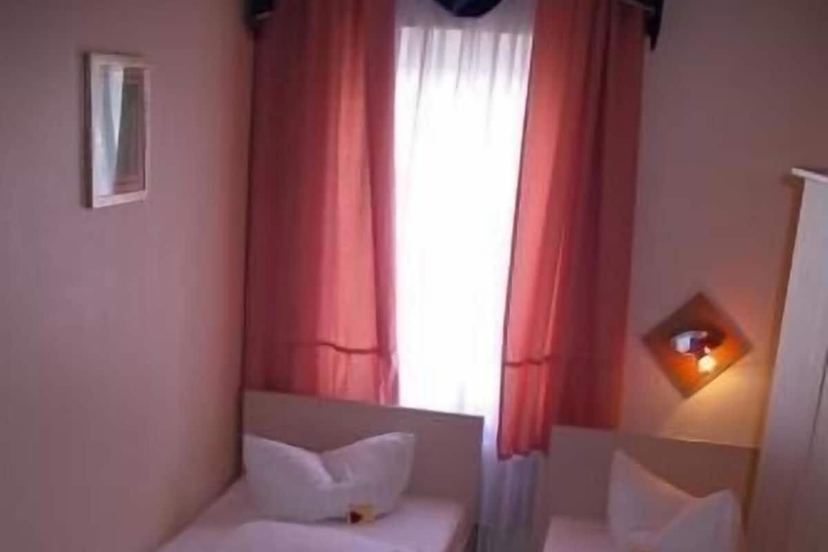 Une petite chambre d’hôtel, composée d’une seule pièce, avec deux lits, une petite table, une chaise, une fenêtre avec des rideaux et un luminaire fixé au mur.