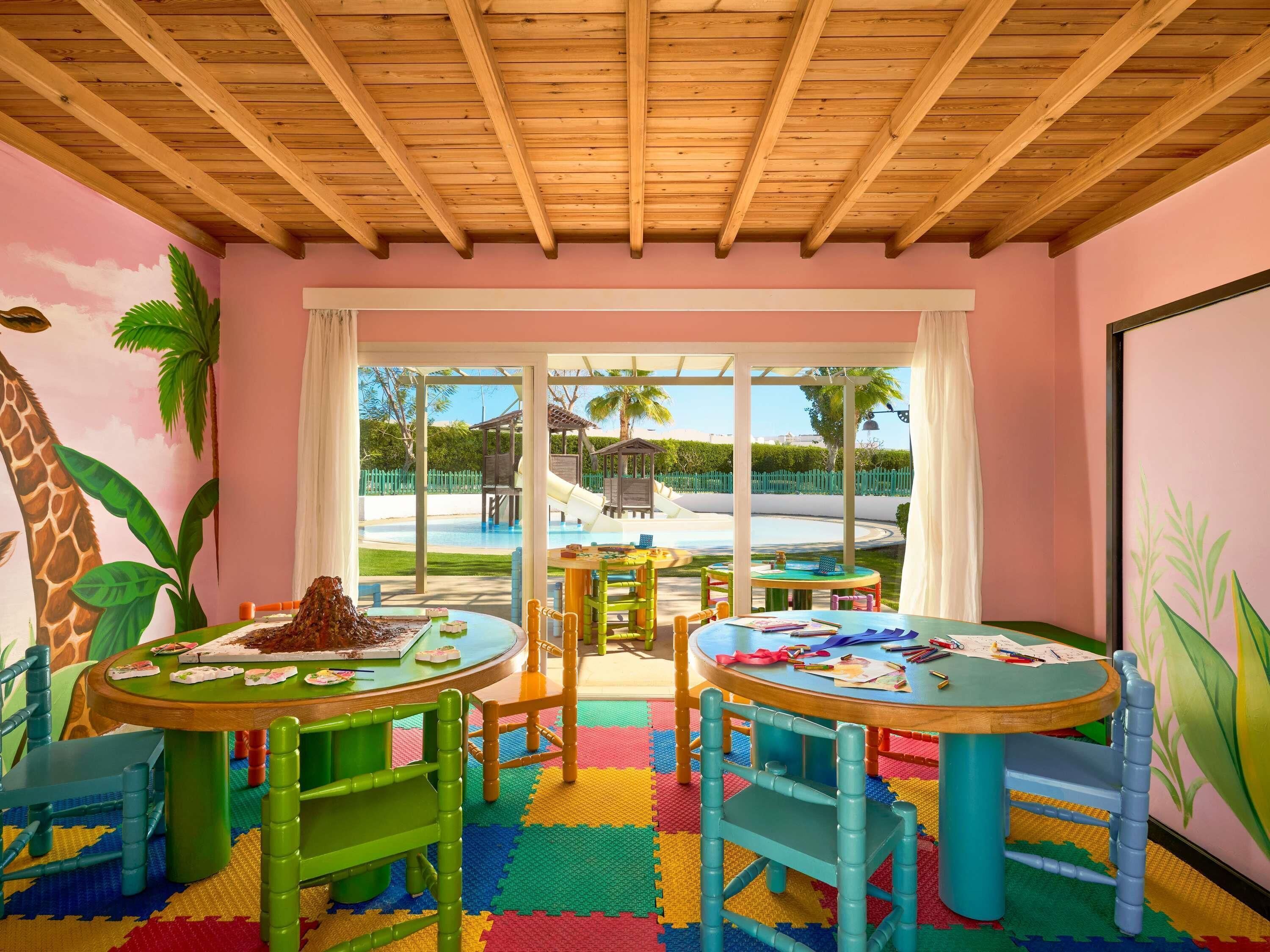 Uma sala de brinquedos infantis com mobiliário colorido e vista para uma piscina e um parquinho.