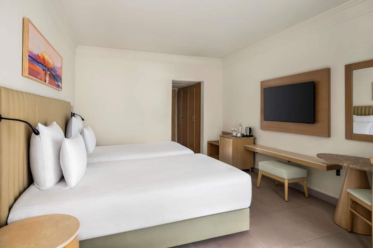 Quarto de hotel com cama, escrivaninha, cadeira, televisão e um quadro na parede.