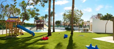 A playground with a slide and climbing structures, surrounded by palm trees and a pool.