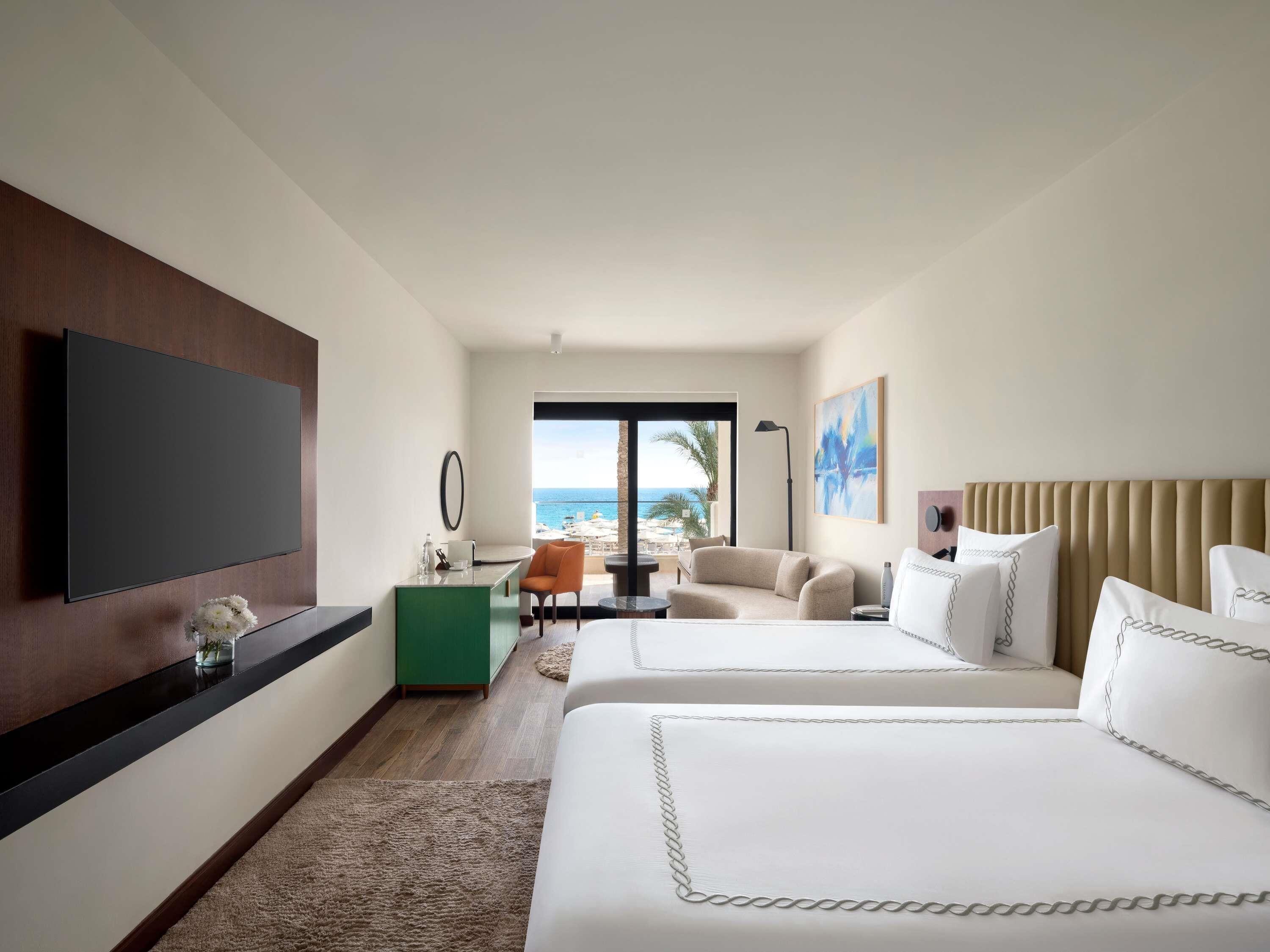 A hotel room with a large TV, a desk with a chair, two beds, a view of the ocean, and a painting on the wall.
