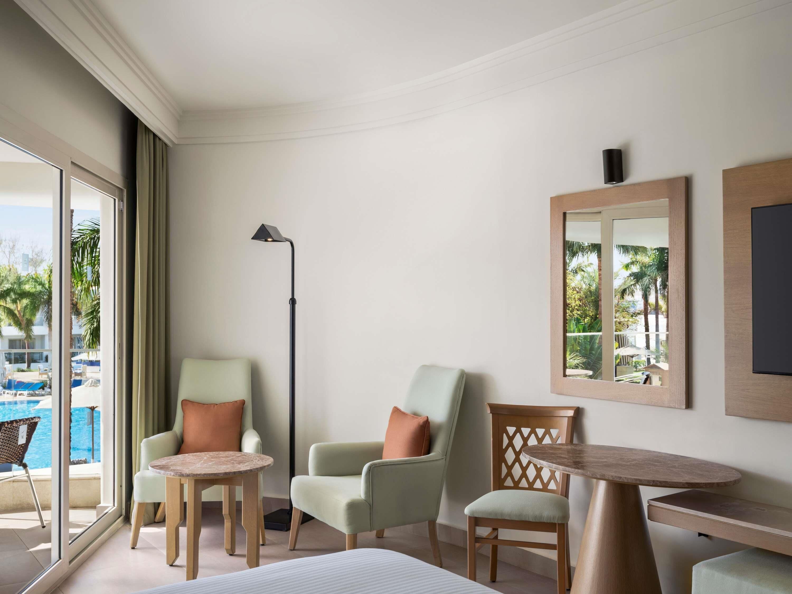 Een hotelkamer met een groot raam, twee fauteuils, een vloerlamp, een kleine tafel en een spiegel aan de muur.