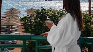 A person in a white robe holding a cup, overlooking a beach with umbrellas and palm trees. - Novotel Sharm El-Sheikh (Sharm El Sheikh)