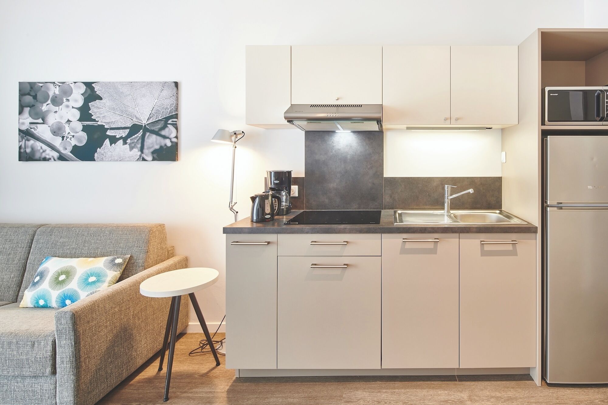A compact kitchen with a built-in stove, a microwave, and a refrigerator. There is a small round table and a sofa with a cushion.