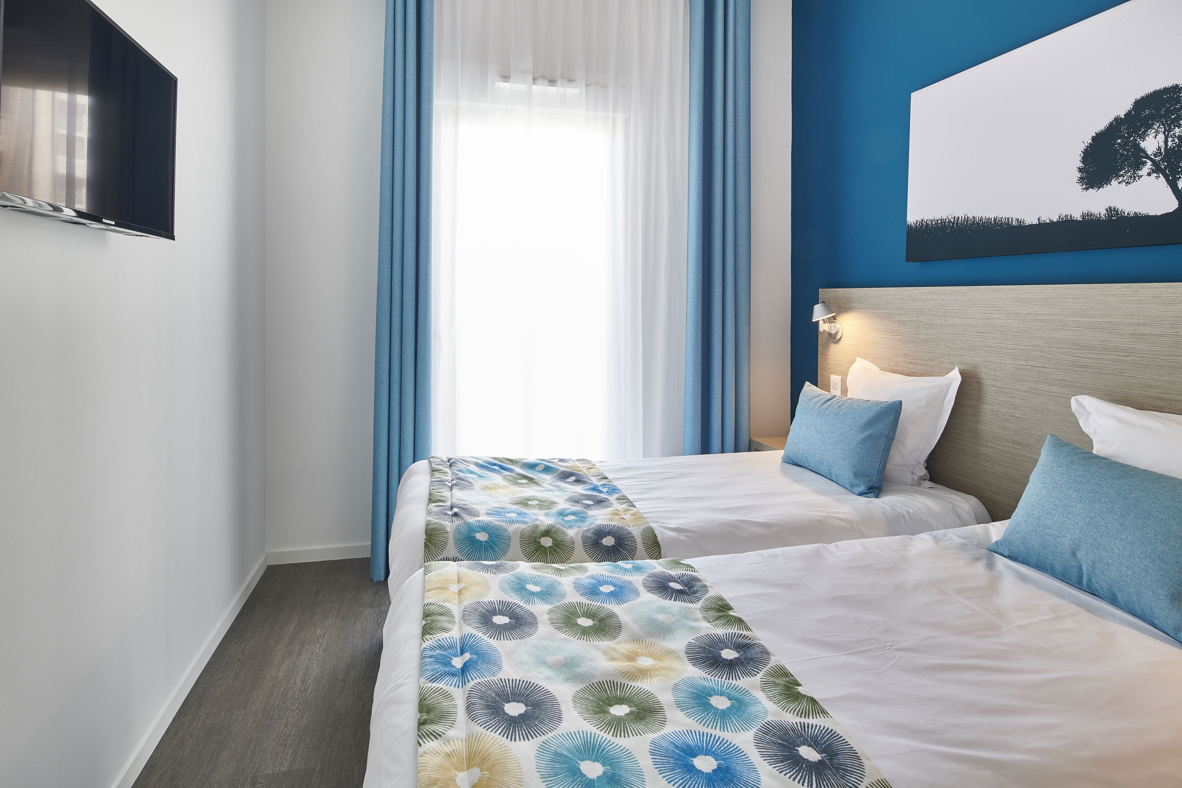 A hotel room with two beds, a large window with blue curtains, a flat-screen TV mounted on the wall, and a painting of a tree on the wall.