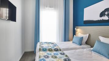 Quarto de hotel com duas camas, uma janela grande com cortinas azuis, uma televisão de tela plana na parede e um quadro de uma árvore na parede.