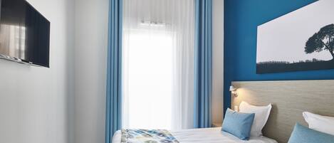 A hotel room with two beds, a large window with blue curtains, a flat-screen TV mounted on the wall, and a painting of a tree on the wall.