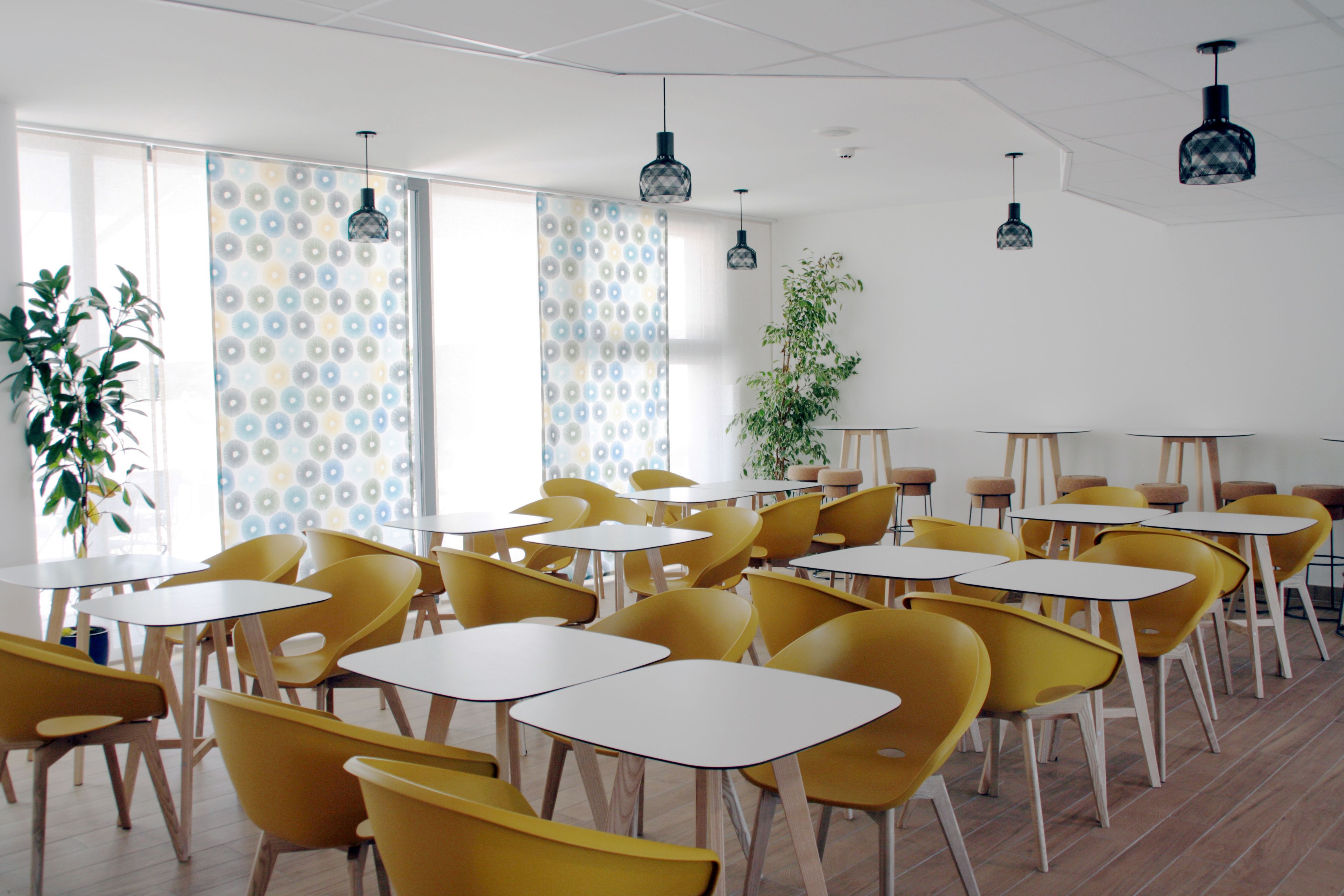 A modern dining area with yellow chairs and white tables, patterned curtains, and hanging pendant lights.