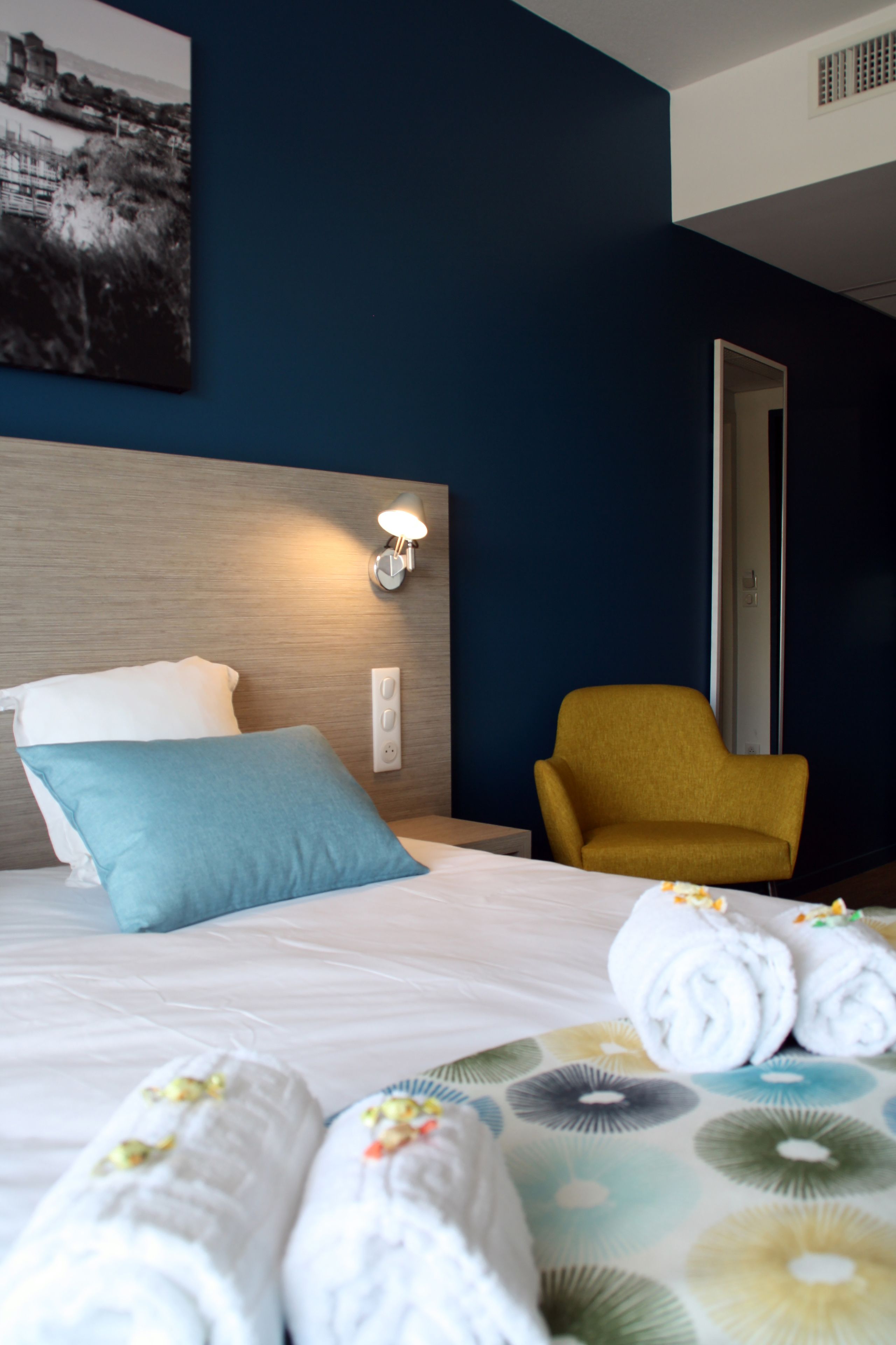 Hotellihuone, jossa on sänky, sängynpääty, seinään kiinnitetty valaisin, keltainen nojatuoli ja seinällä oleva kehystetty kuva.