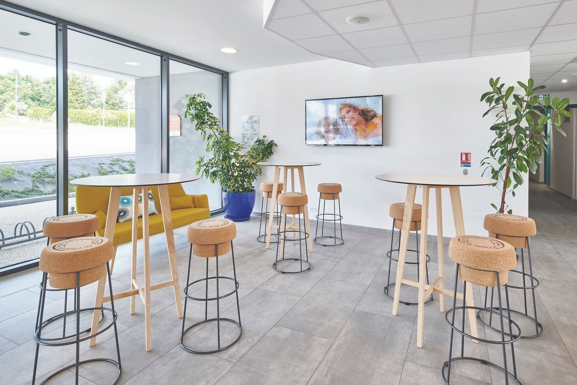 Un intérieur moderne avec des tables et des tabourets ronds, de grandes fenêtres et des plantes d’intérieur.