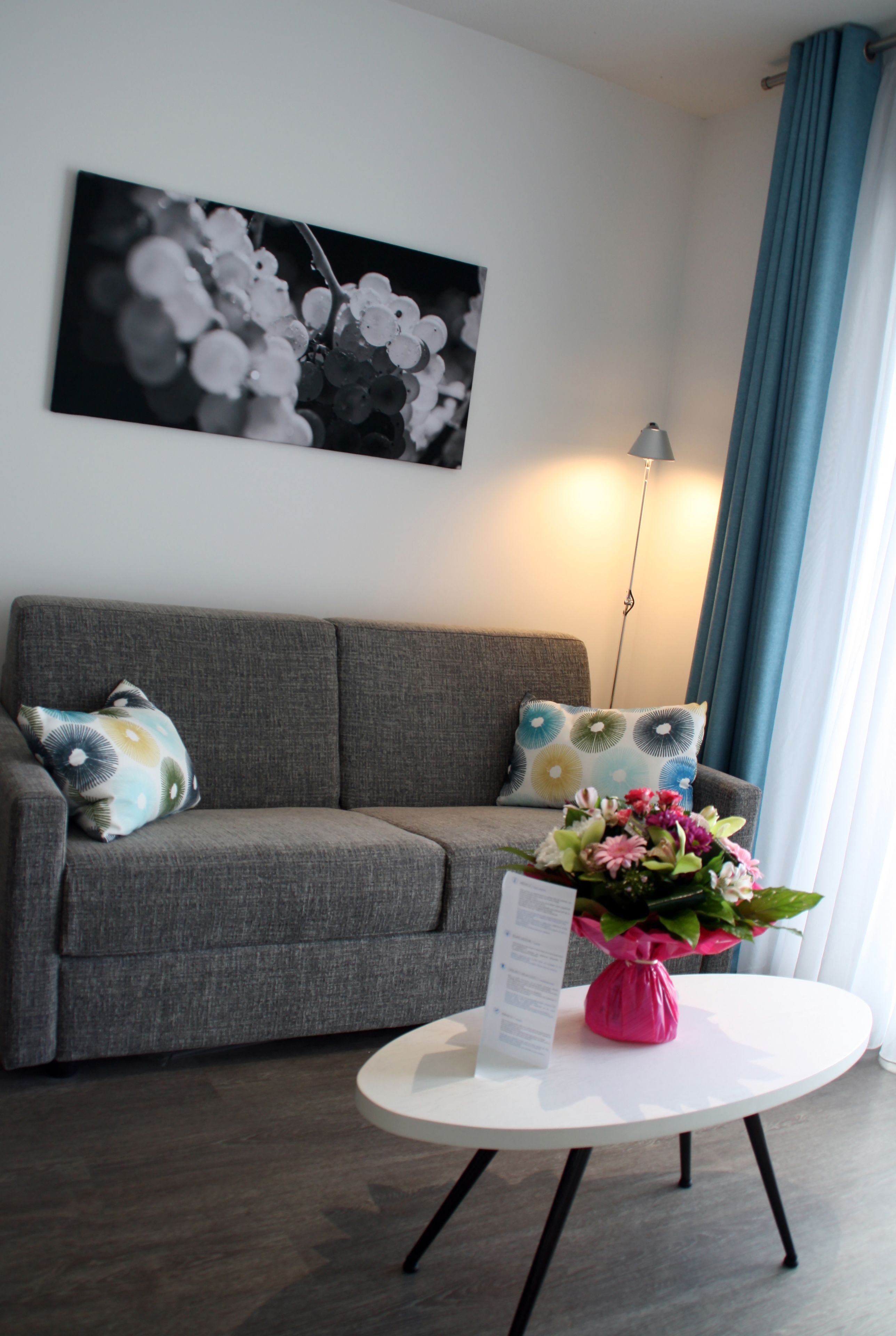 A modern living room with a grey sofa, patterned cushions, a white coffee table with a pink vase, and a black and white artwork on the wall.
