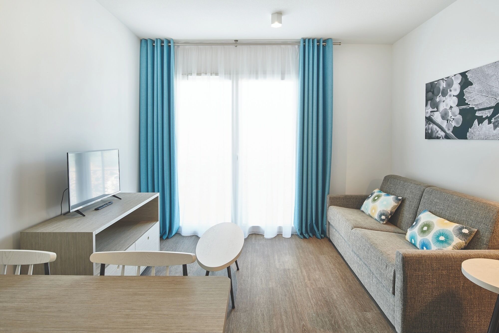Ein modernes Wohnzimmer mit Fernseher, einer Couch mit gemusterten Kissen, einem Esstisch und einem großen Fenster mit blauen Vorhängen.