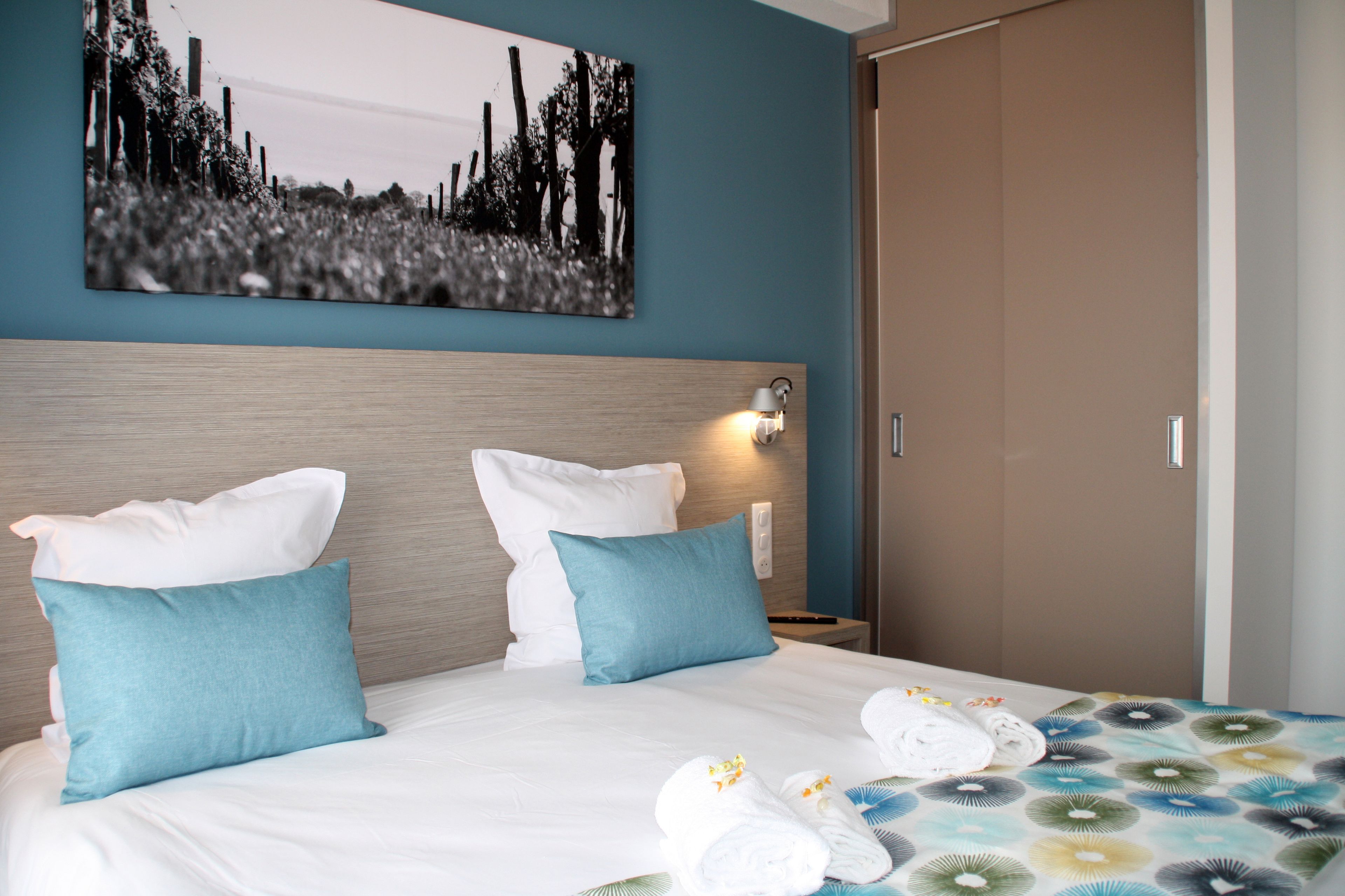 A hotel room with a bed, two pillows, a nightstand, a wall-mounted lamp, and a door.
