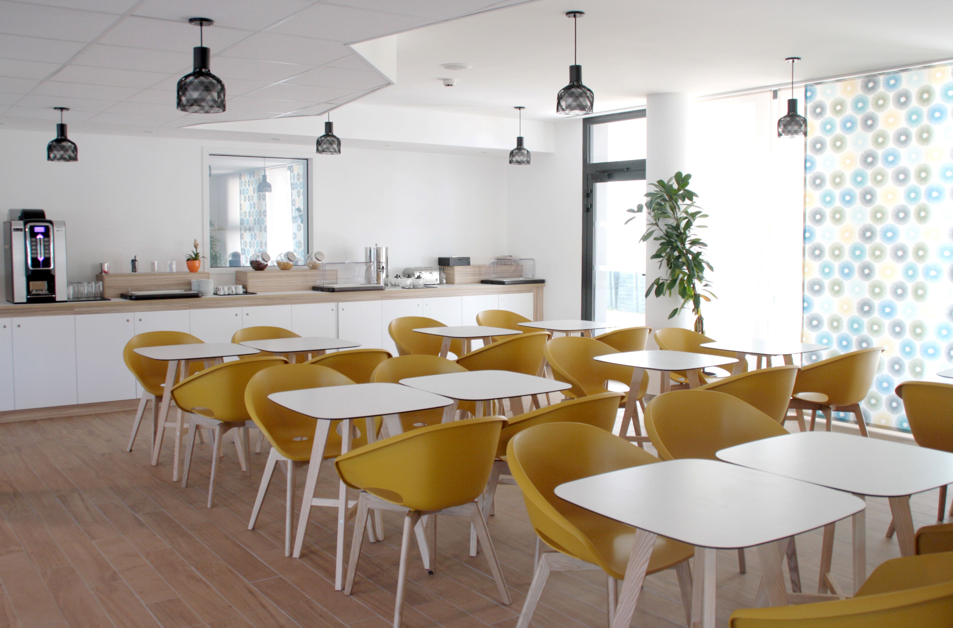 Uma sala de jantar moderna com cadeiras amarelas e mesas brancas, uma máquina de café e uma janela grande com uma planta.