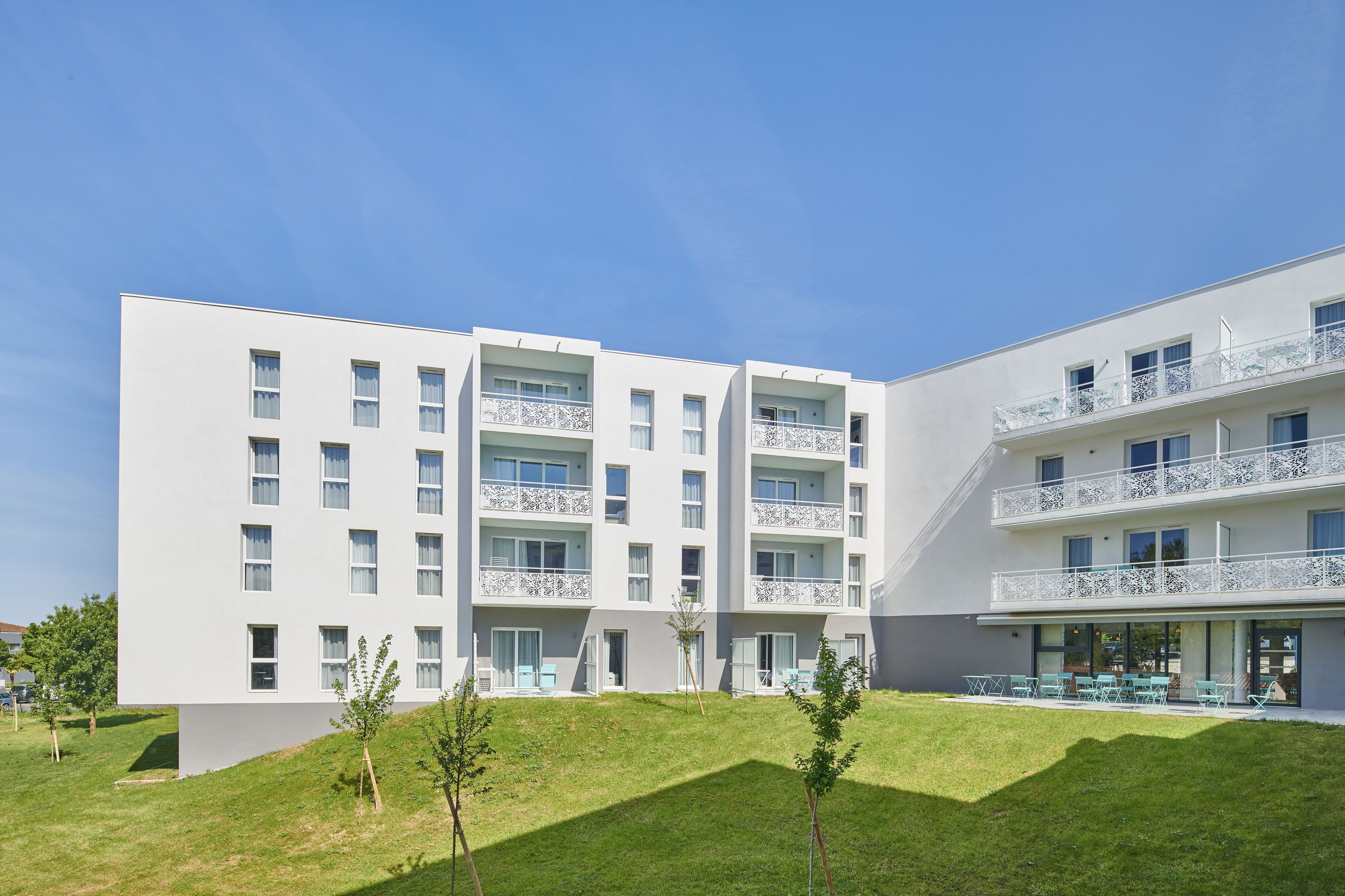 Un edificio moderno de varios pisos con balcones, techo plano y una zona verde con árboles pequeños.