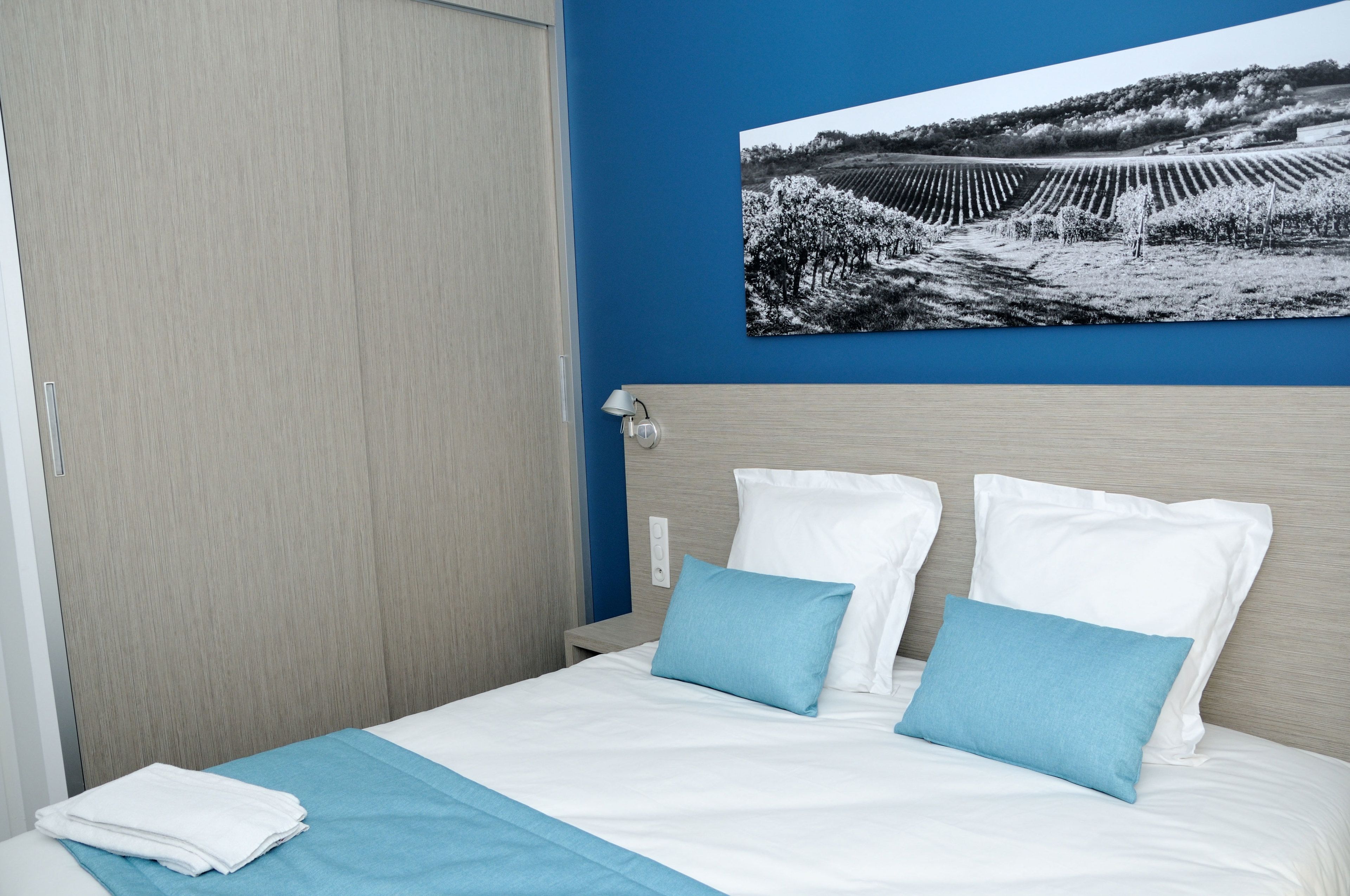A neatly made bed with white and blue bedding, a blue wall, and a black and white photograph of a vineyard.