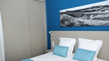 A neatly made bed with white and blue bedding, a blue wall, and a black and white photograph of a vineyard.