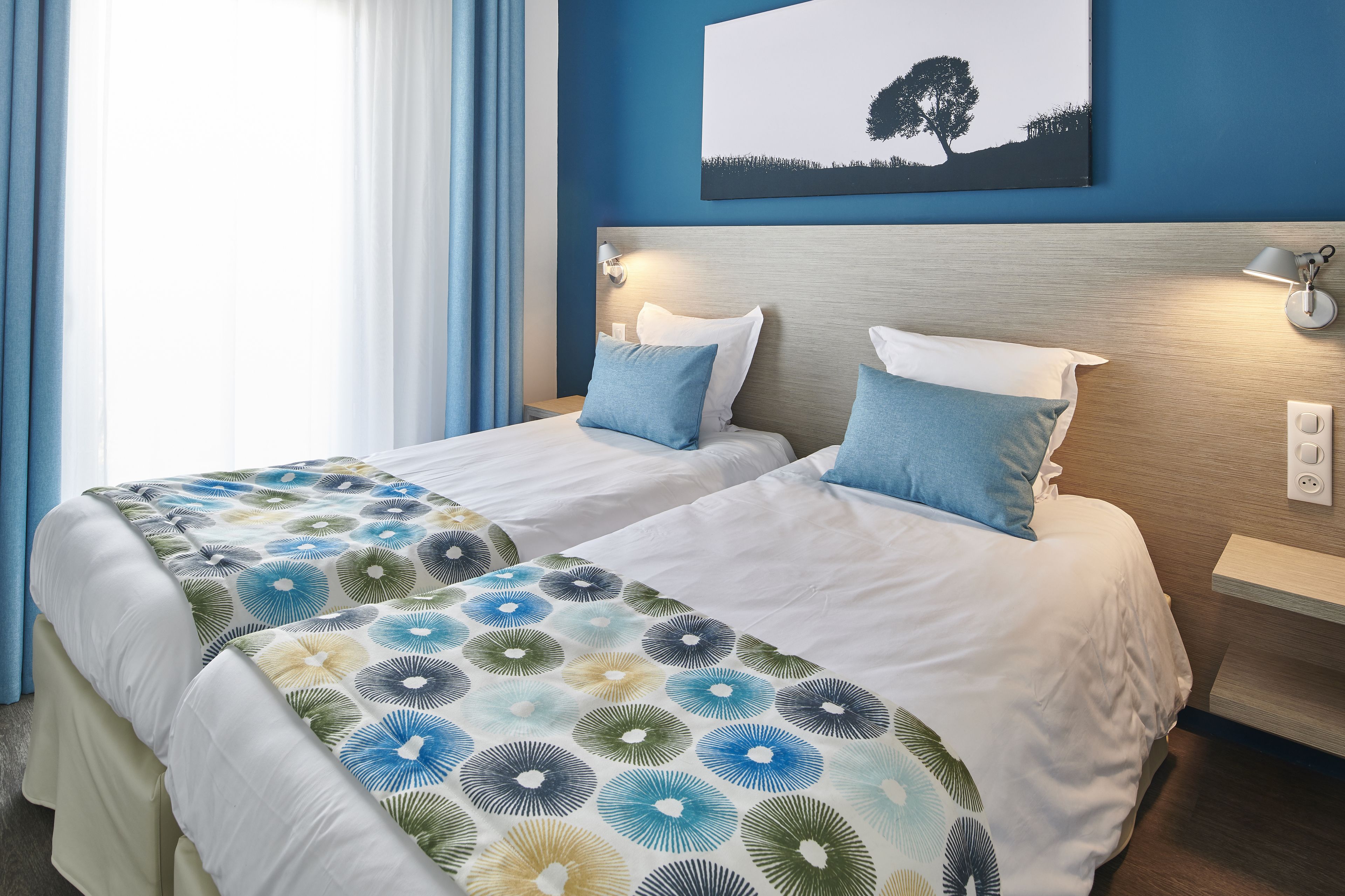 Une chambre d’hôtel avec deux lits, une table de chevet, une lampe fixée au mur et un tableau représentant un arbre.