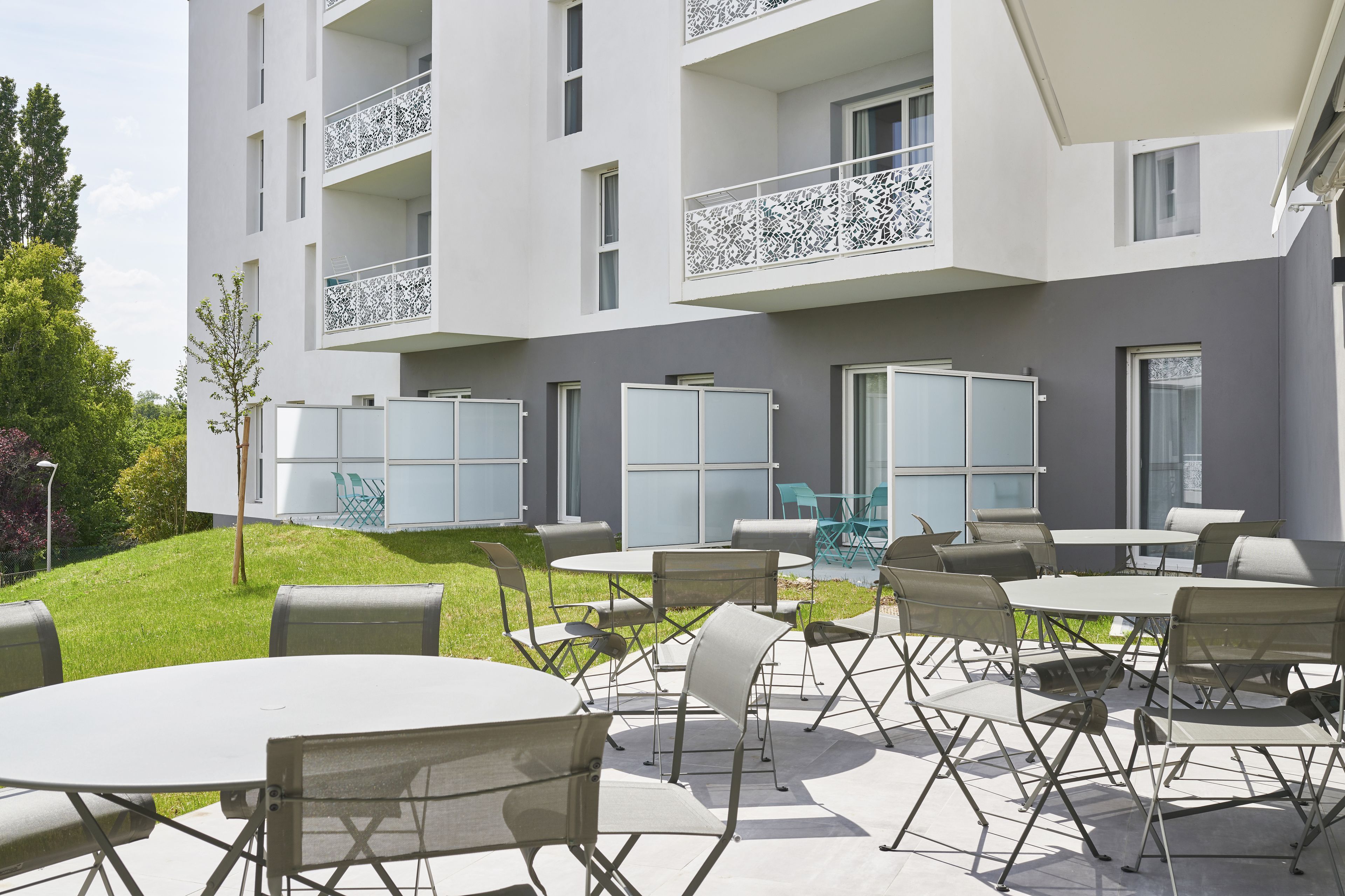 A modern building with multiple balconies, outdoor seating area with tables and chairs, and a well-maintained lawn.
