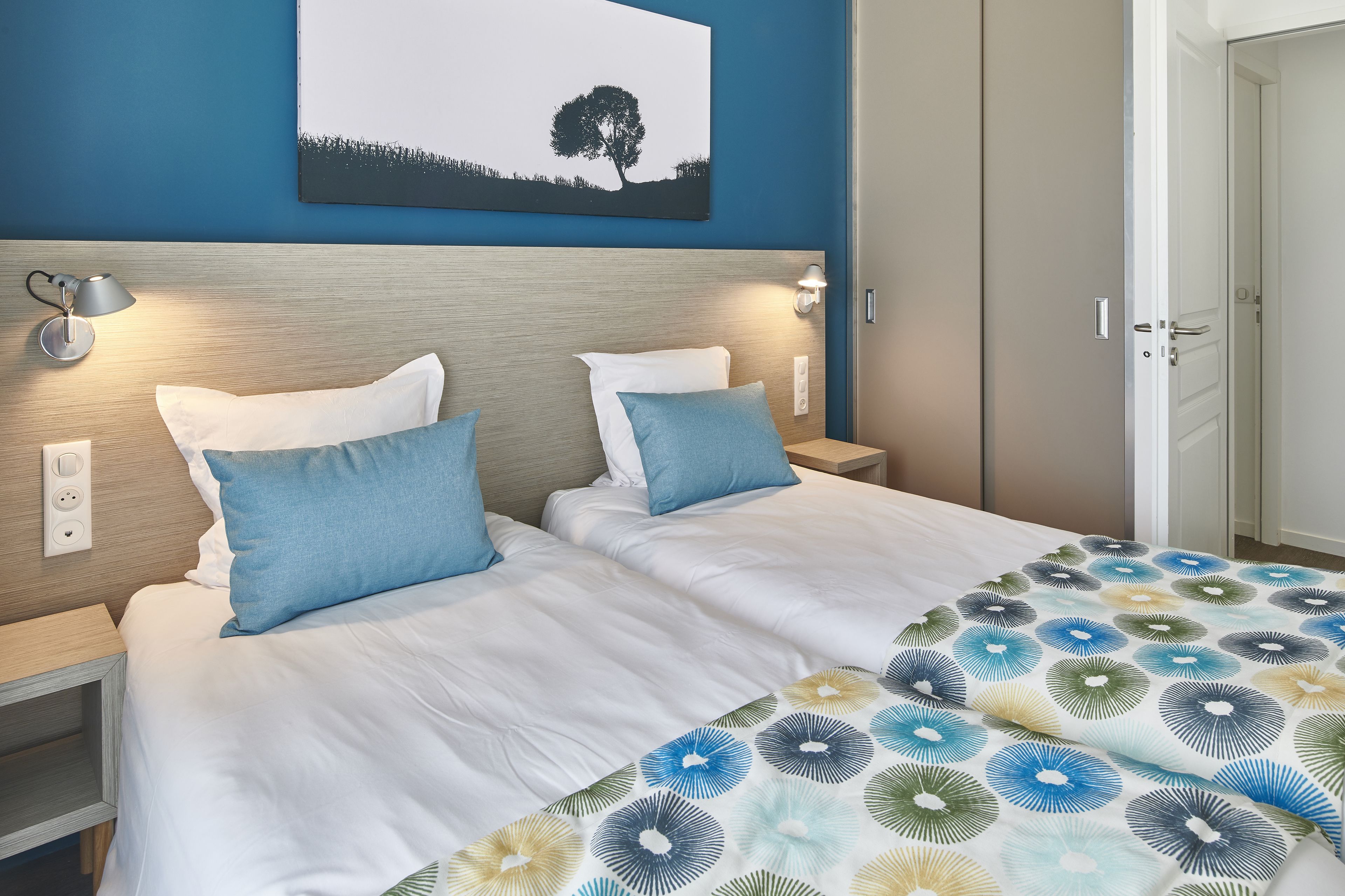 Cama doble con ropa de cama blanca y almohadas azules, una mesita de noche con lámpara, y un cuadro colgado en la pared de un árbol.