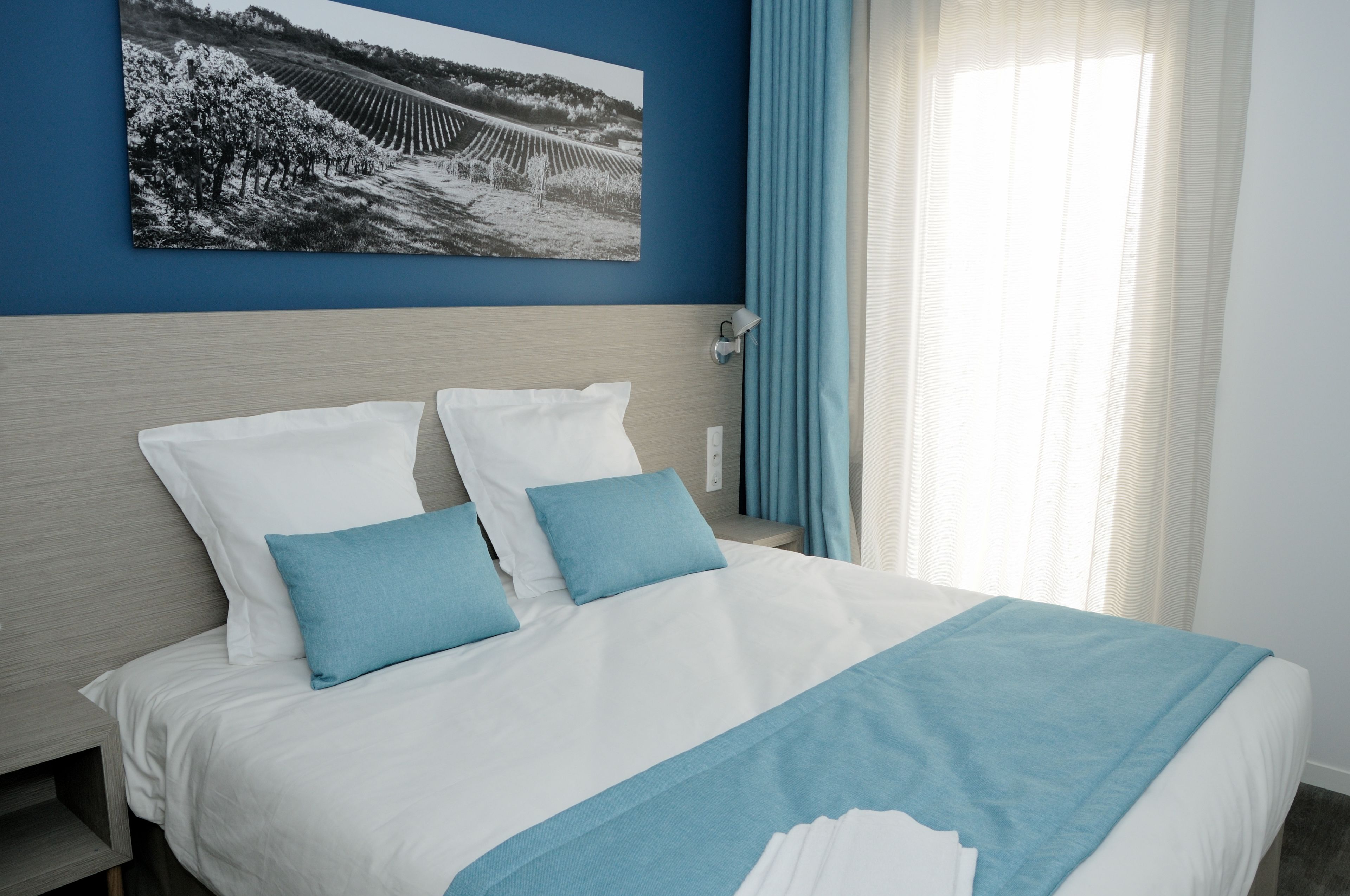 Una cama bien tendida con ropa de cama blanca y azul, un cabecero y un cuadro colgado en la pared que muestra un viñedo.