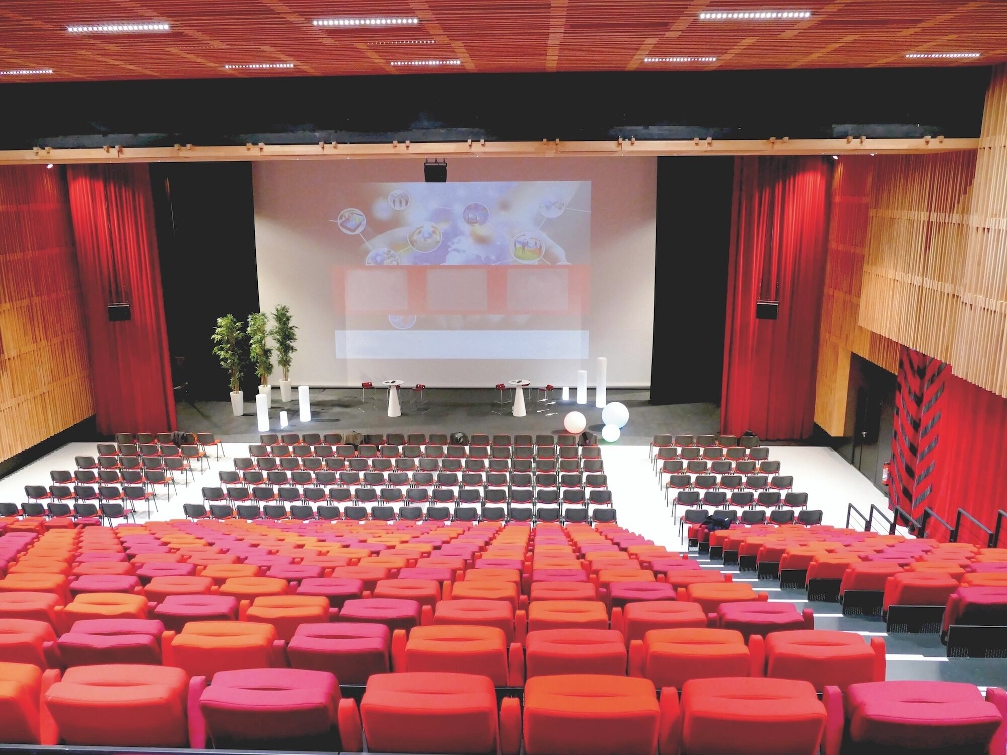 An auditorium with rows of red and orange seats, a stage with a large screen, and red curtains.