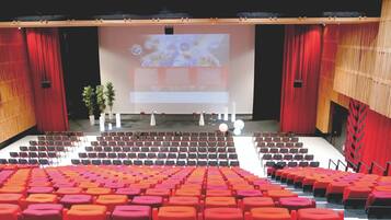 An auditorium with rows of red and orange seats, a stage with a large screen, and red curtains.