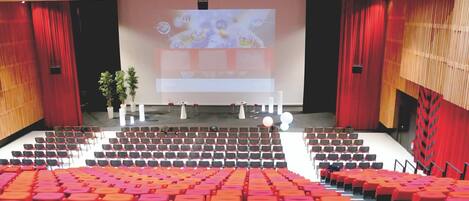 Un auditorio con hileras de asientos rojos y anaranjados, un escenario con una pantalla grande y cortinas rojas.