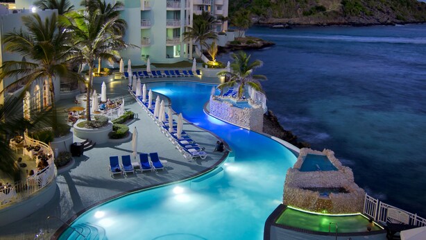 A resort pool area with a curved design, lounge chairs, umbrellas, and a building with a light-colored facade.