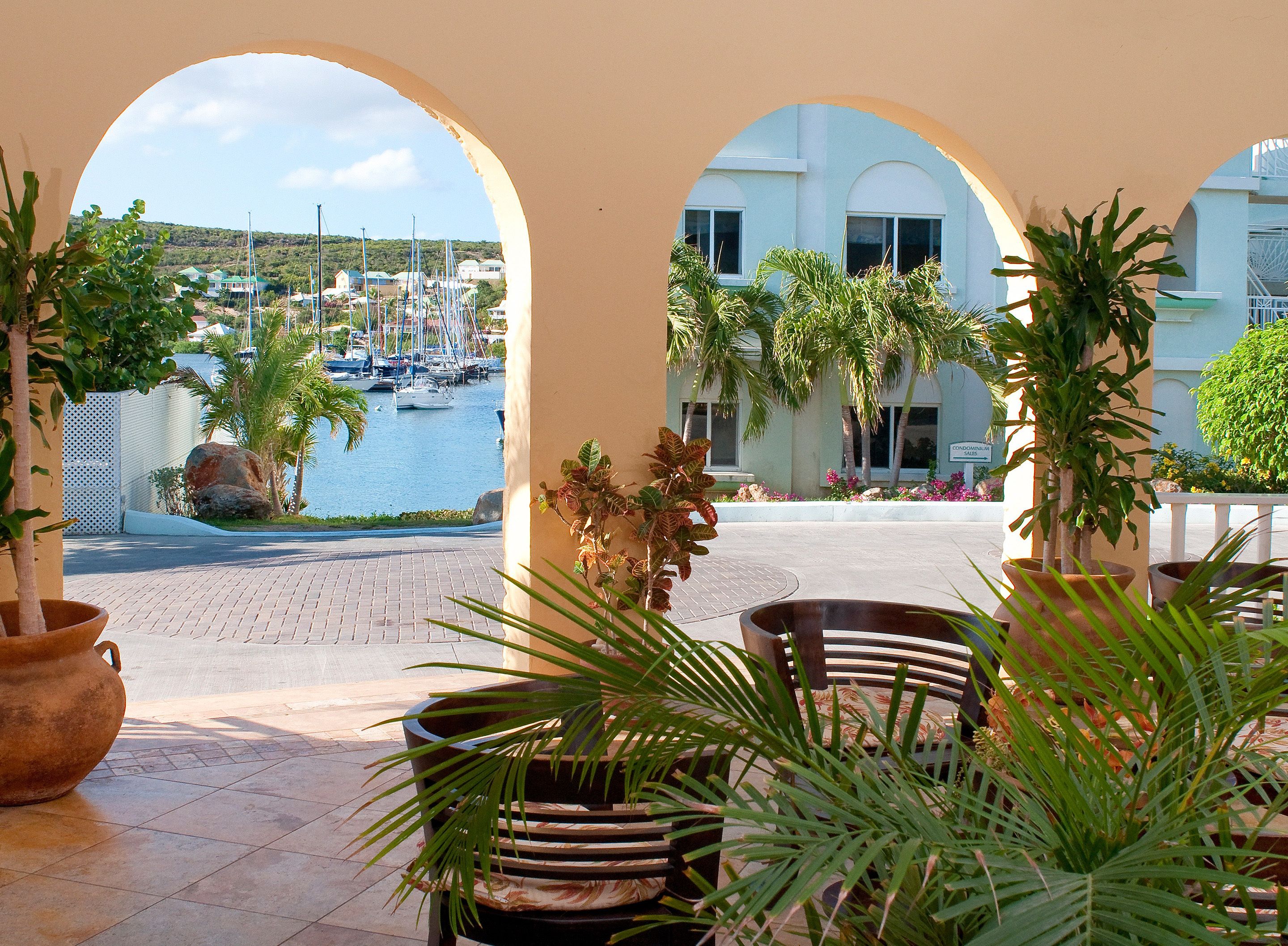 A view through an archway of a marina with boats, a building with arched windows, and potted plants.
