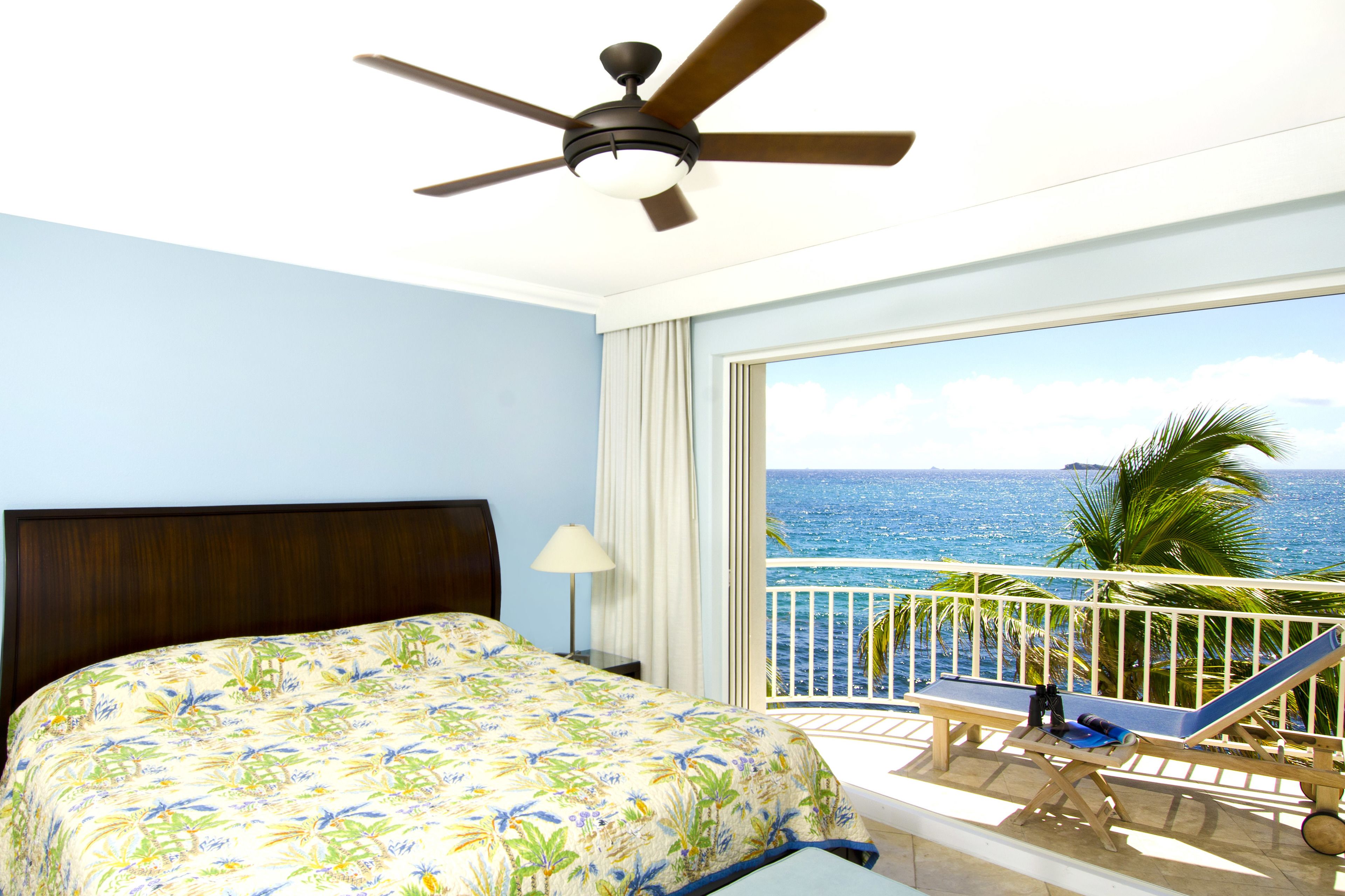A hotel room with a bed, a ceiling fan, a balcony with a view of the ocean, and a chair.