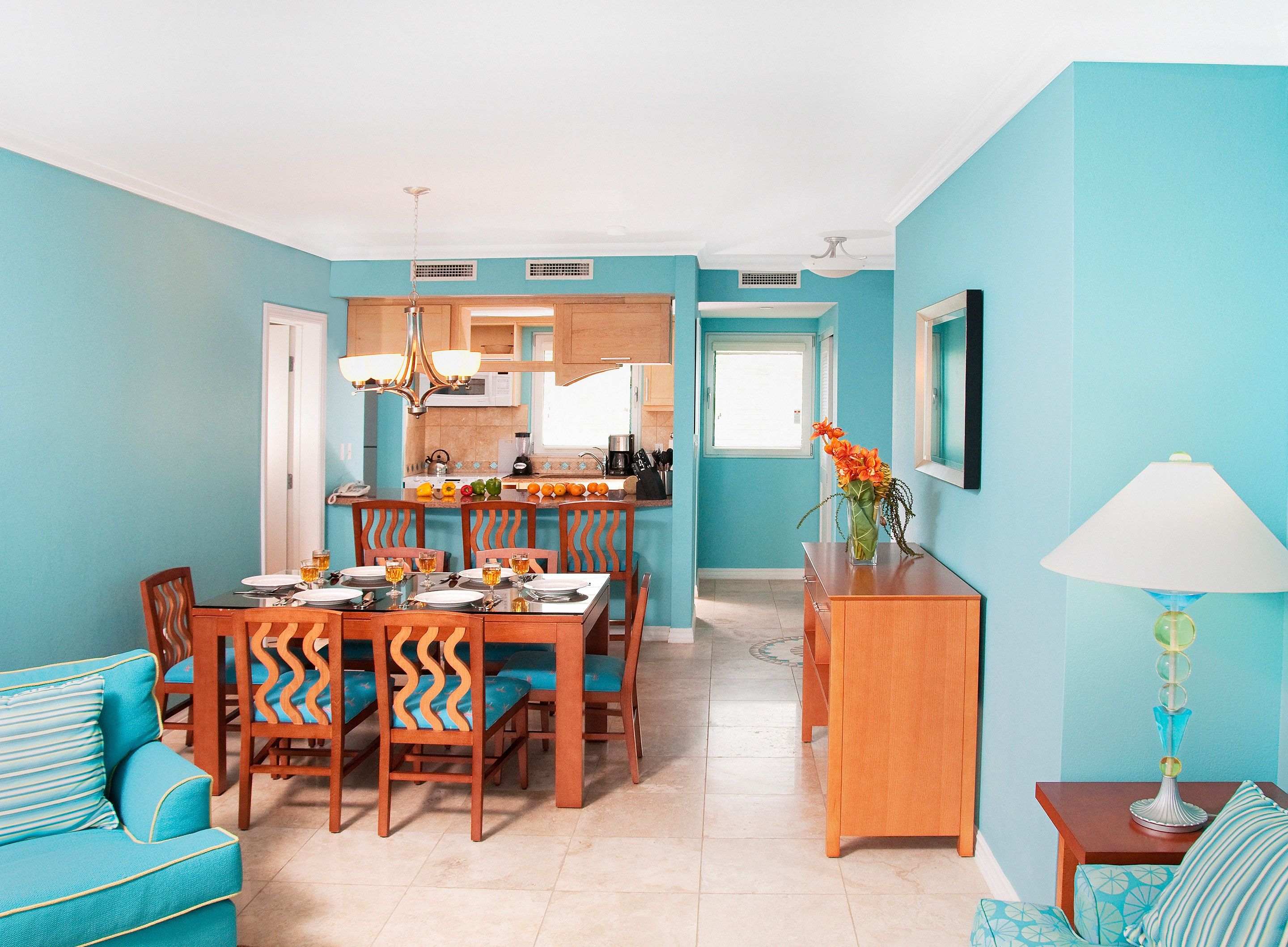 A dining area with a wooden table and chairs, a kitchen in the background, and a teal-colored living room with a sofa and a lamp.