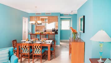 A dining area with a wooden table and chairs, a kitchen in the background, and a teal-colored living room with a sofa and a lamp.