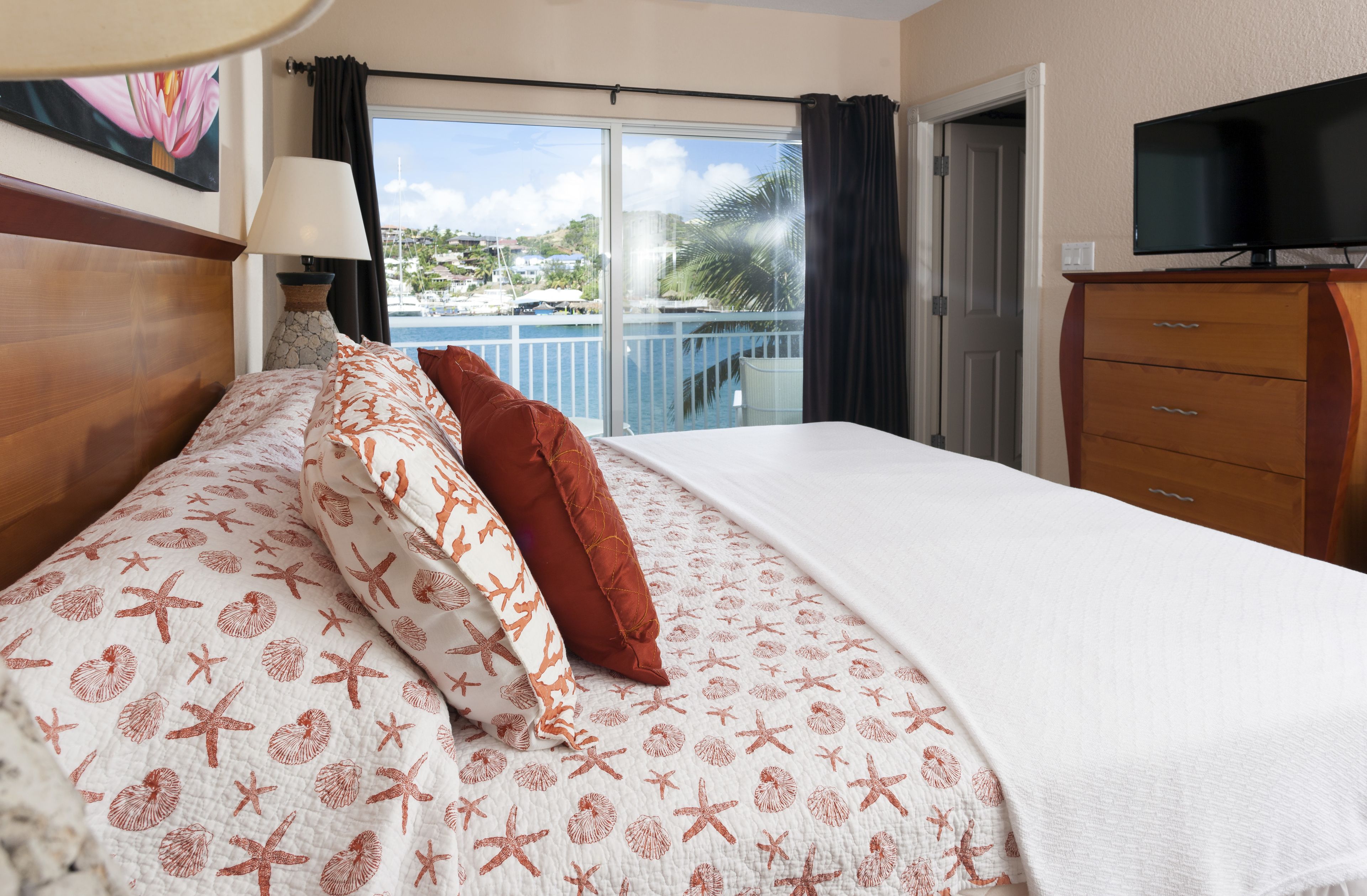 A hotel room with a bed, a TV, a dresser, and a balcony with a view of the sea and buildings.