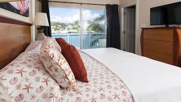 A hotel room with a bed, a TV, a dresser, and a balcony with a view of the sea and buildings.