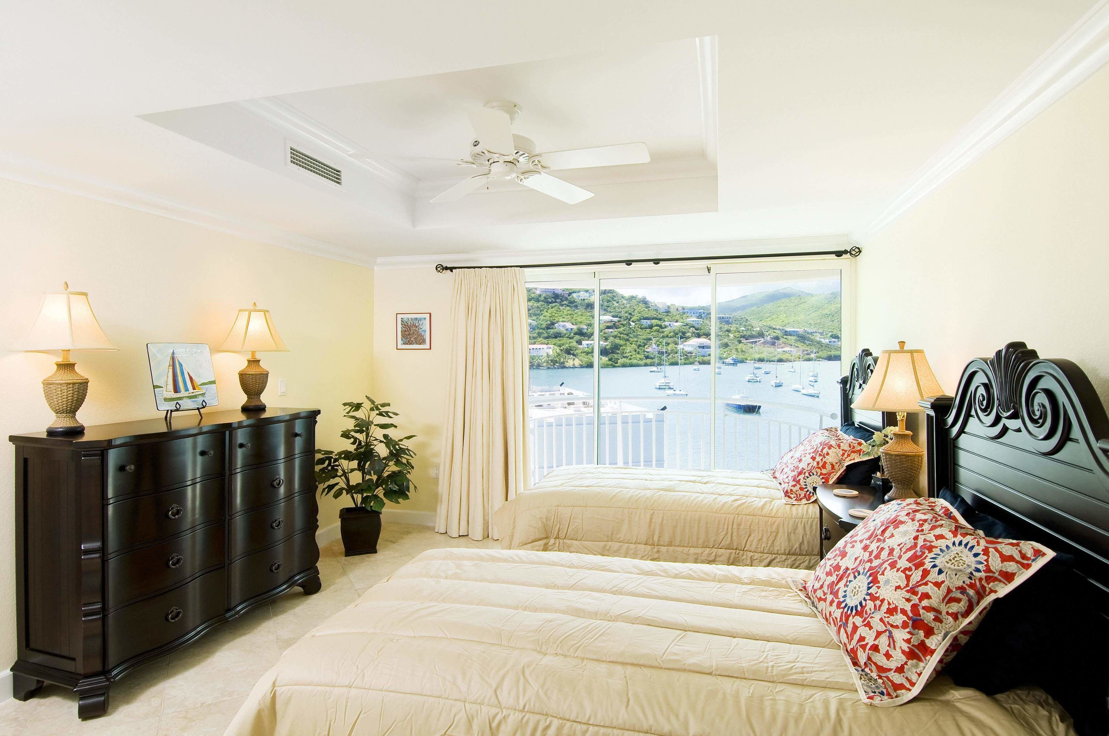 A bedroom with two beds, a dark wooden dresser, a ceiling fan, and a large window with a view of boats and hills.