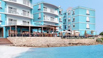 A multi-story hotel with balconies, a restaurant area, and a stone wall by the water.