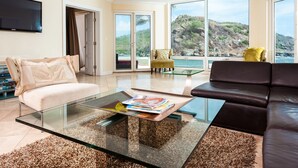 A modern living room with a glass coffee table, a beige sofa, a black leather sofa, and a view of a mountainous landscape through large windows.