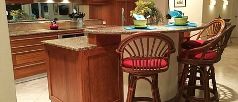 A kitchen with wooden cabinets, a stainless steel range hood, a dishwasher, and a bar area with two wicker barstools.