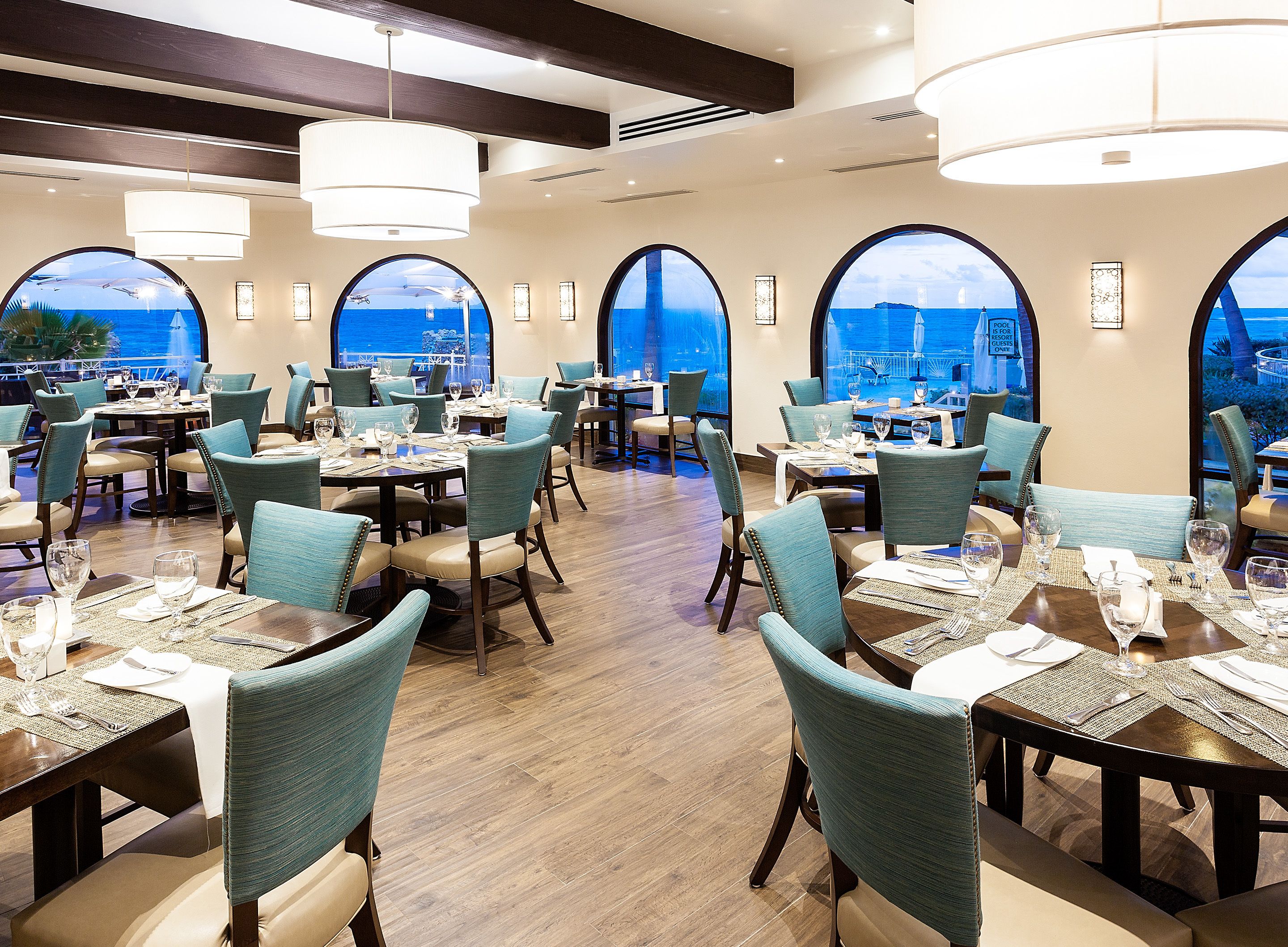 Restaurante com mesas redondas, cadeiras verde-água e grandes janelas com vista para o oceano.