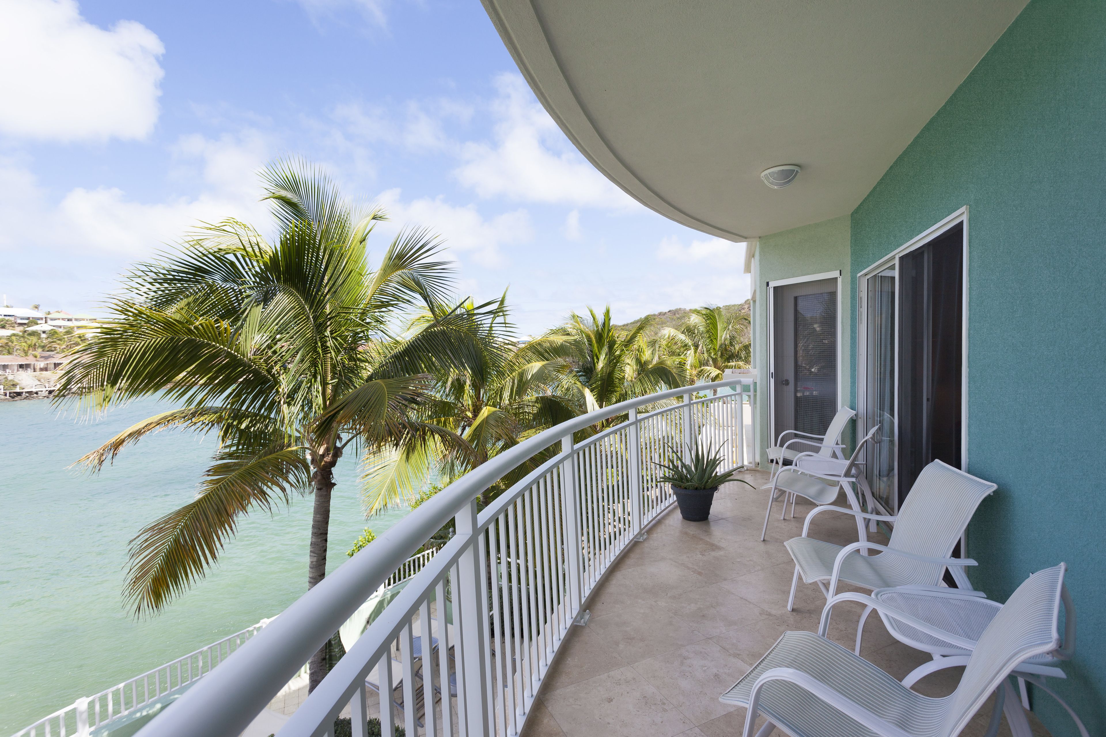 A balcony with white chairs and a potted plant, overlooking a body of water and palm trees.