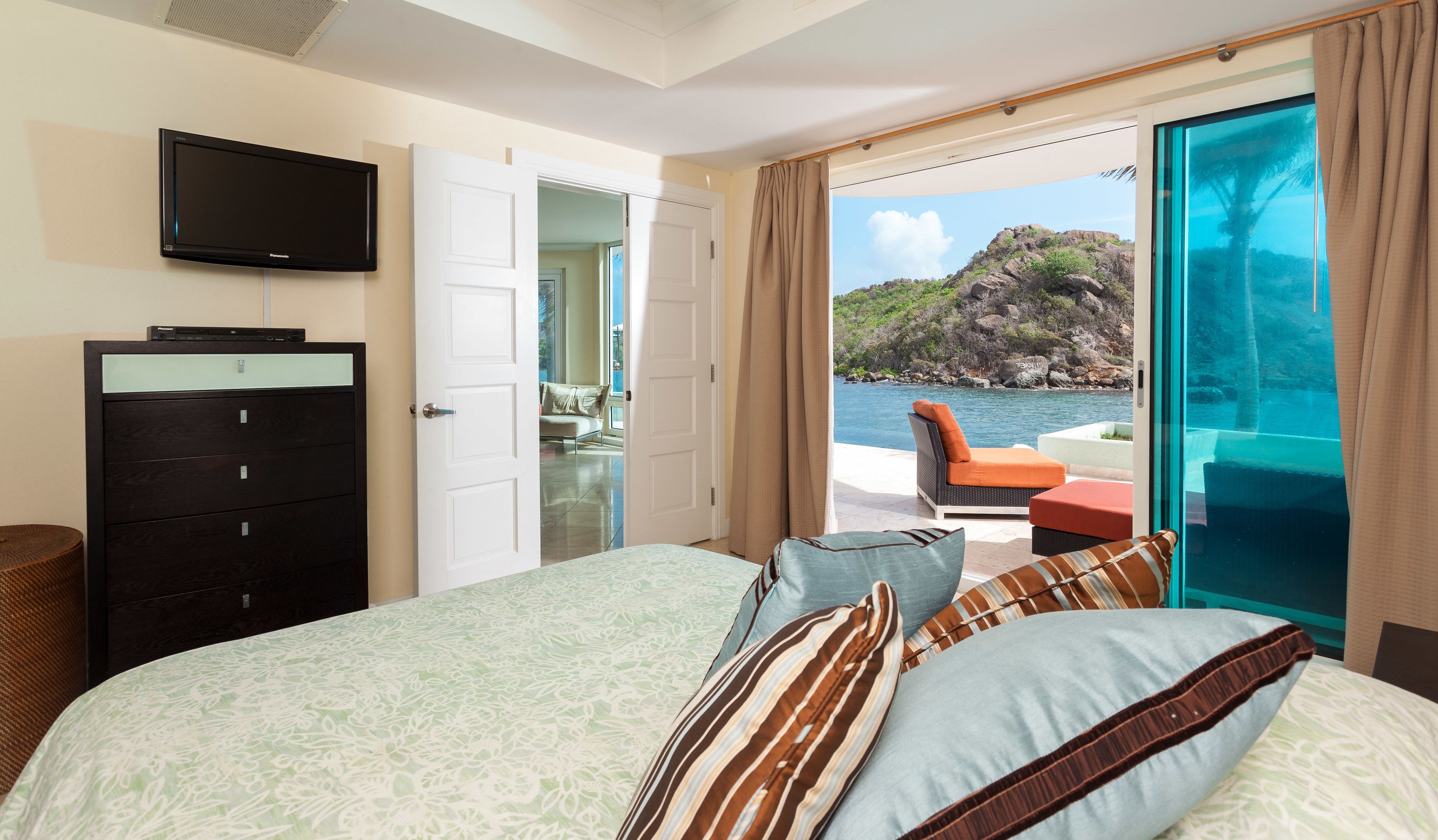 A bedroom with a bed, a TV, a dresser, and a view of the sea.