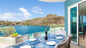 A dining table set for six with a bottle of wine, surrounded by wicker chairs, overlooking a body of water and a mountainous landscape.