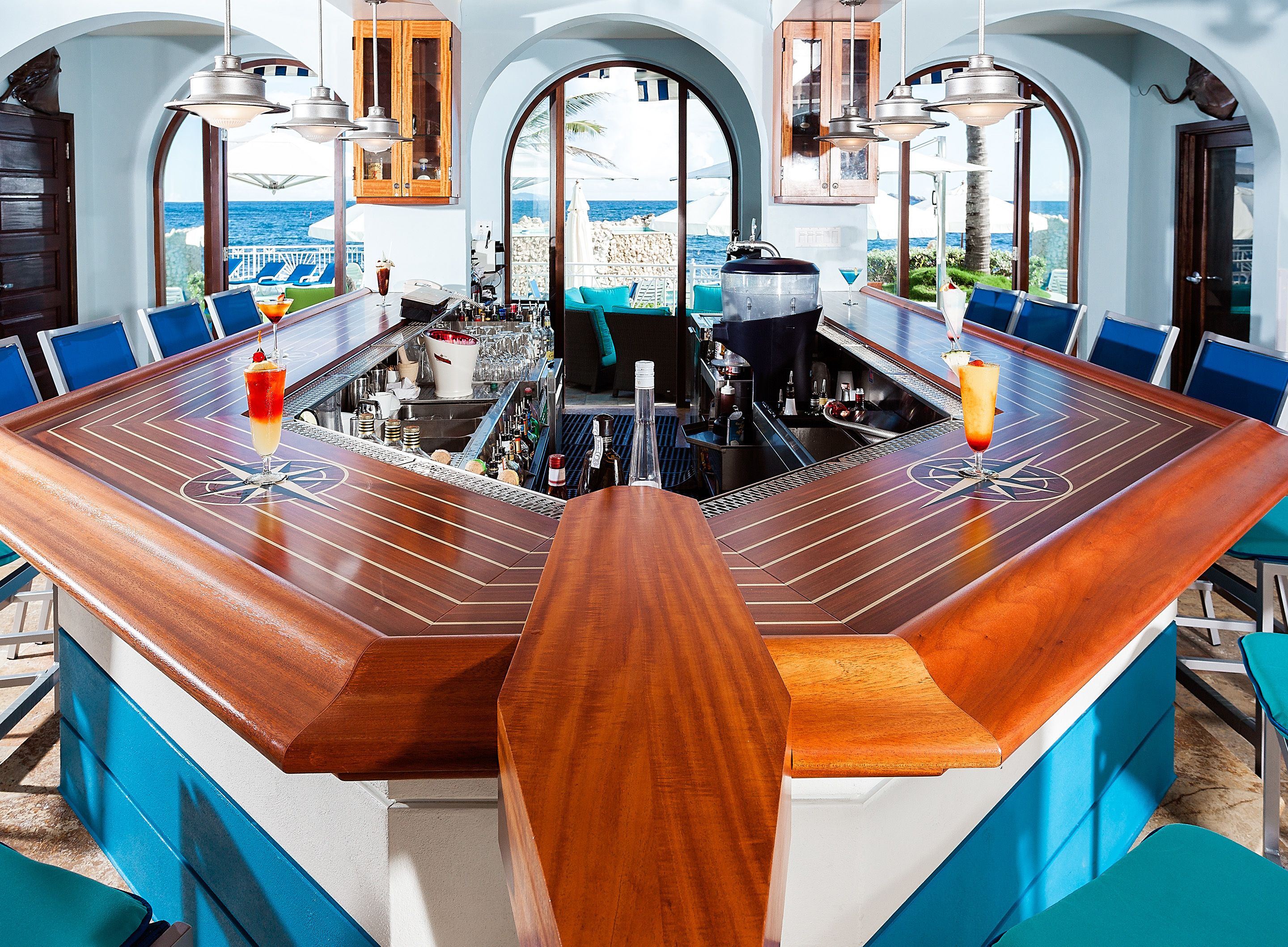 Zona de bar con mostrador de madera, sillas azules y vista al océano.