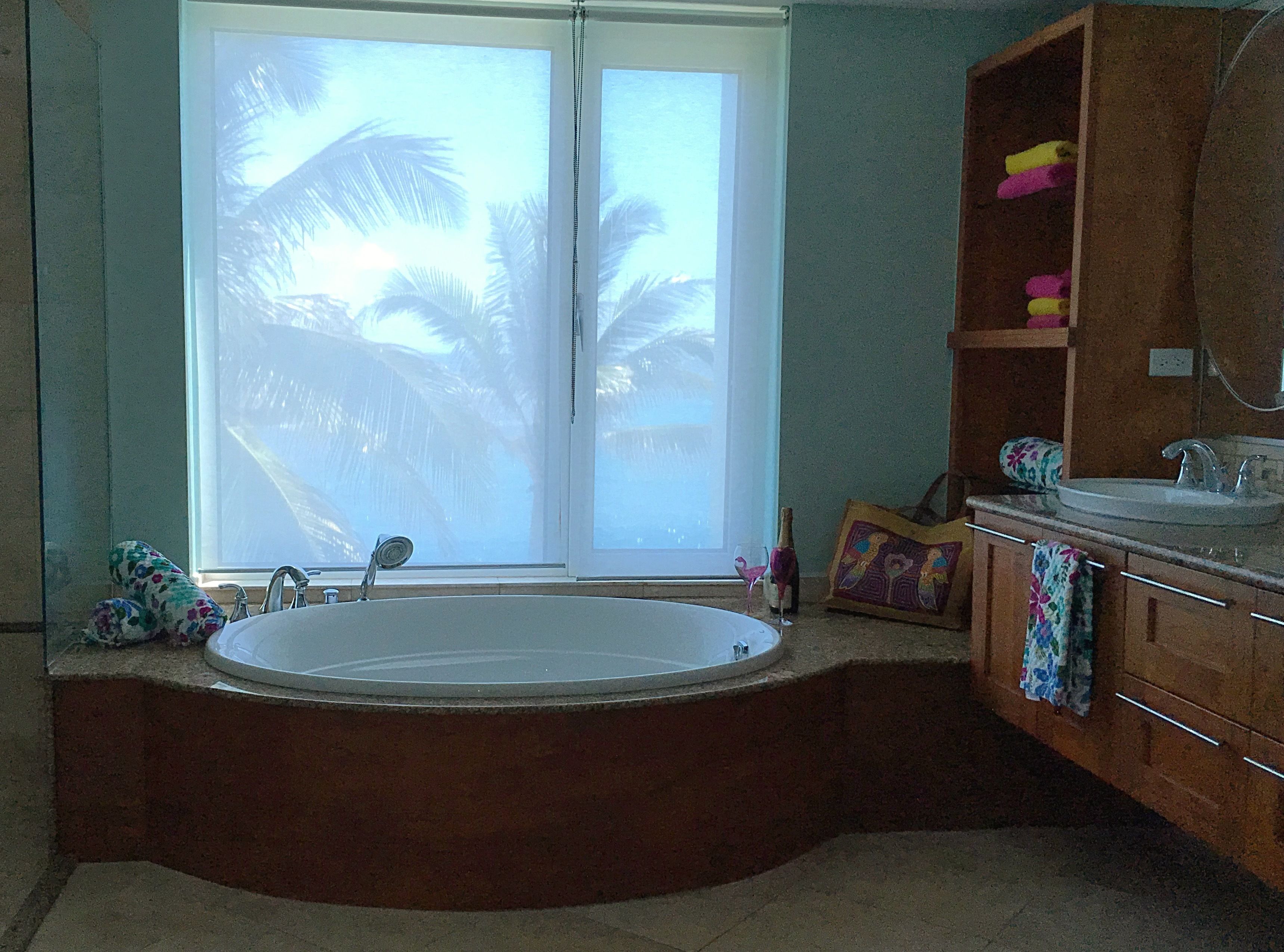 A bathroom with a large bathtub, a window with a view of palm trees, and a wooden vanity with a mirror.
