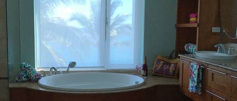 A bathroom with a large bathtub, a window with a view of palm trees, and a wooden vanity with a mirror.