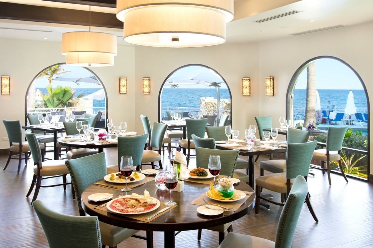 Un restaurante con mesas redondas, sillas verdes y amplios ventanales con vistas al mar.
