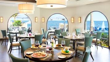 Un restaurante con mesas redondas, sillas verdes y amplios ventanales con vistas al mar.